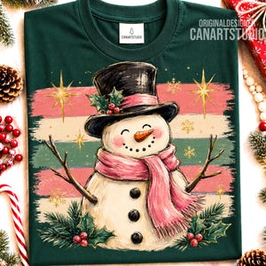 Może przedstawiać: Ciemnozielony t-shirt z grafiką bałwana w czarnym cylindrze z ostrokrzewem i r&oacute;żowym szalikiem. Wz&oacute;r zawiera poziome pociągnięcia pędzla w kolorze r&oacute;żowym, kremowym i zielonym, złote gwiazdki oraz płatki śniegu na ciemnozielonym tle.