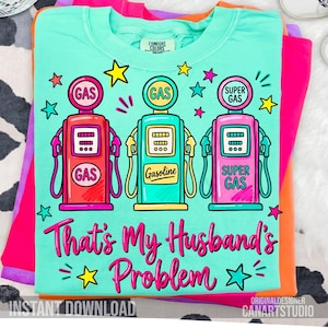 Könnte beinhalten: Türkisfarbenes T-Shirt mit einer Grafik von drei Retro-Zapfsäulen in Rot, Türkis und Pink, mit dem Text "That's My Husband's Problem". Das T-Shirt ist auf andere farbige T-Shirts gefaltet.