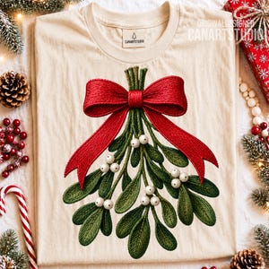 Könnte beinhalten: Ein cremefarbenes T-Shirt mit einer Stickerei, die einen grünen Mistelzweig mit weißen Beeren und einer großen roten Schleife zeigt. Das Etikett am Innenkragen trägt den Text CANARTSTUDIO mit einem kleinen Baum-Symbol.