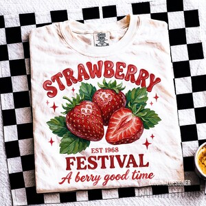 May include: Cream-coloured t-shirt with a strawberry design. The design features the word "STRAWBERRY" in red, three strawberries, green leaves, and the text "EST 1968 FESTIVAL A berry good time."