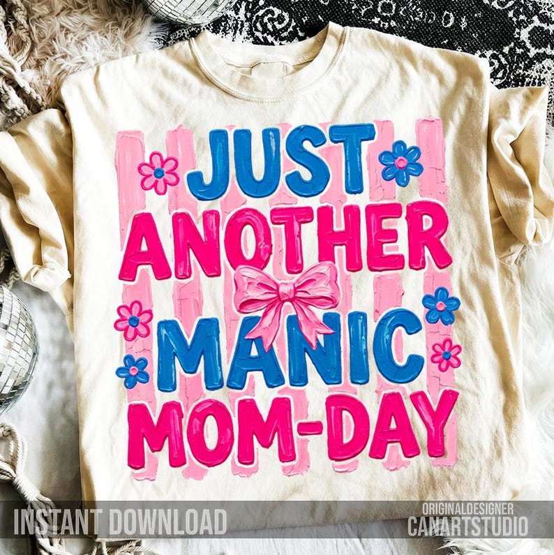 Op de afbeelding: Cr&egrave;mekleurig T-shirt met de tekst "JUST ANOTHER MANIC MOM-DAY" in blauw en roze. Het ontwerp bevat roze verticale strepen, bloemen en een strik. De woorden "INSTANT DOWNLOAD" staan onderaan.