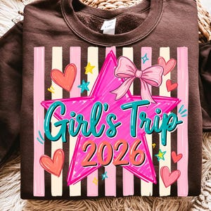 May include: A brown sweatshirt with a graphic design. The design features a pink star with the words "Girl's Trip 2026" in teal and orange, a pink bow, hearts, and stars. Vertical stripes in pink and white are in the background.