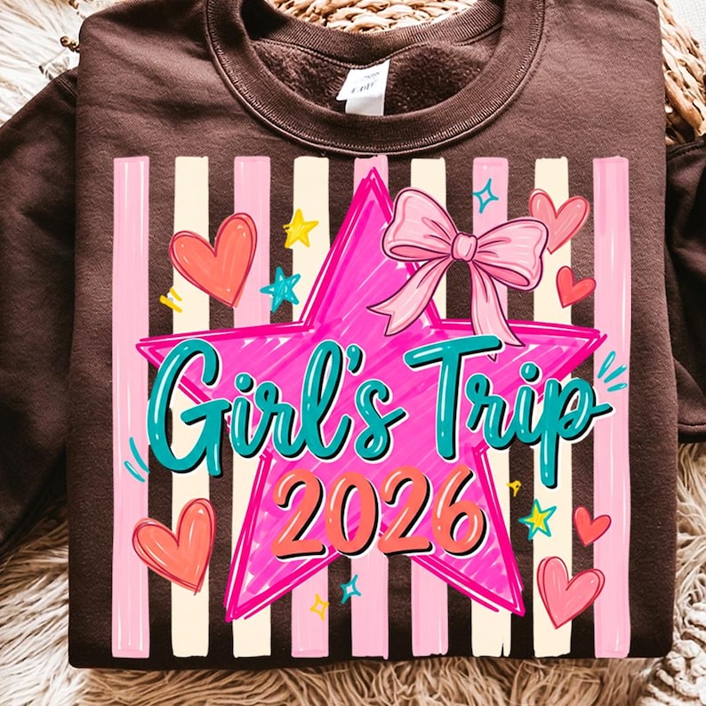 May include: A brown sweatshirt with a graphic design. The design features a pink star with the words "Girl's Trip 2026" in teal and orange, a pink bow, hearts, and stars. Vertical stripes in pink and white are in the background.