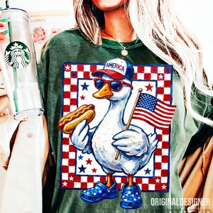 May include: Green t-shirt featuring a patriotic duck wearing sunglasses and a baseball cap that says "America." The duck holds a hot dog and an American flag. The design is set against a red and white checkered background with stars.
