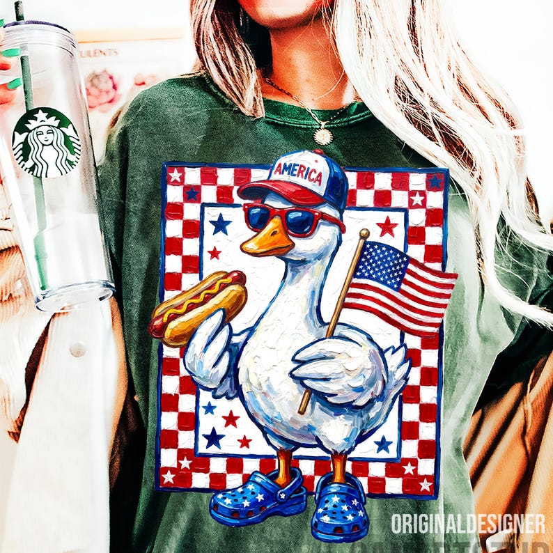 May include: Green t-shirt featuring a patriotic duck wearing sunglasses and a baseball cap that says "America." The duck holds a hot dog and an American flag. The design is set against a red and white checkered background with stars.