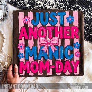 Op de afbeelding: Een bruin t-shirt met de tekst "JUST ANOTHER MANIC MOM-DAY" in blauw en roze, versierd met roze bloemen en een roze strik. De woorden staan tegen roze verticale strepen.