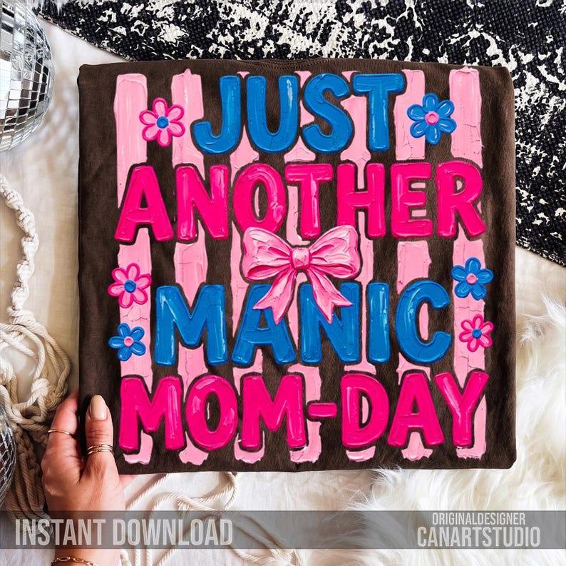Op de afbeelding: Een bruin t-shirt met de tekst "JUST ANOTHER MANIC MOM-DAY" in blauw en roze, versierd met roze bloemen en een roze strik. De woorden staan tegen roze verticale strepen.