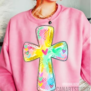 Op de afbeelding: Roze sweatshirt met een kleurrijk kruisontwerp. Het kruis is versierd met aquarel spetters in geel, groen, roze en blauw, geaccentueerd met goudglitter en kleine hartjes. De tekst "ORIGINALDESIGNER CANARTSTUDIO" is zichtbaar.