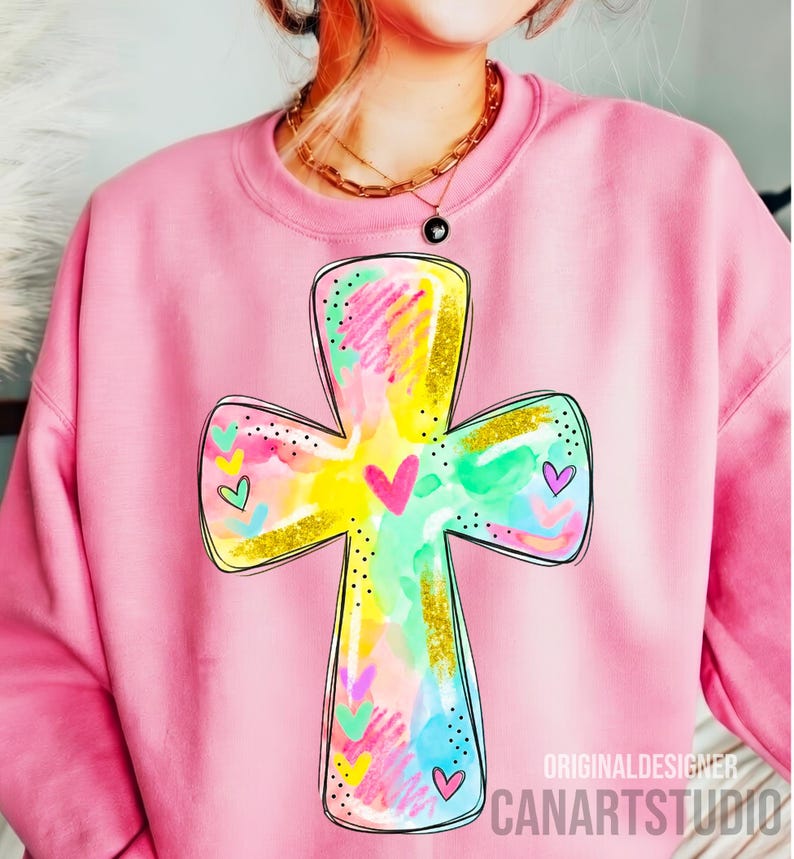 Op de afbeelding: Roze sweatshirt met een kleurrijk kruisontwerp. Het kruis is versierd met aquarel spetters in geel, groen, roze en blauw, geaccentueerd met goudglitter en kleine hartjes. De tekst "ORIGINALDESIGNER CANARTSTUDIO" is zichtbaar.