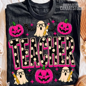 May include: A dark gray t-shirt featuring a graphic print with the word TEACHER in large black letters with white polka dots and a pink outline. The design includes three white ghosts wearing black glasses and holding yellow pencils, three bright pink jack-o'-lanterns, and several small black bats and pink stars.