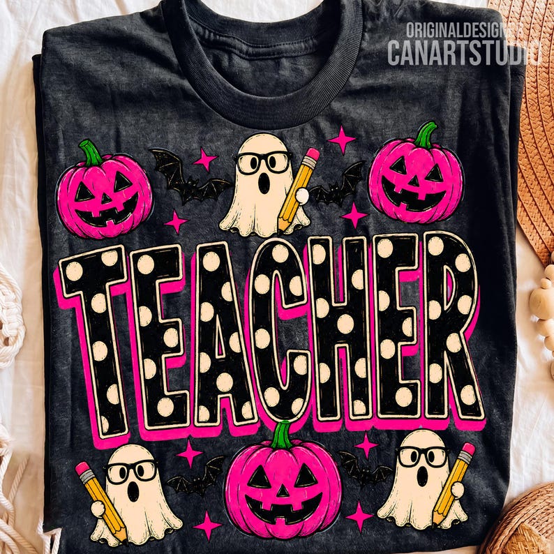 May include: A dark grey t-shirt featuring a graphic print with the word TEACHER in large black letters with white polka dots and a pink outline. The design includes three white ghosts wearing black glasses and holding yellow pencils, three bright pink jack-o'-lanterns, and several small black bats and pink stars.