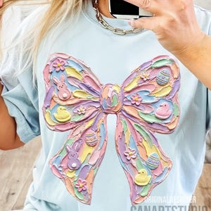 May include: Light blue t-shirt featuring a large, textured bow design in pastel colors. The bow is decorated with Easter-themed elements like bunnies, eggs, and flowers. The text "ORIGINAL DESIGNER CANARTSTUDIO" is visible at the bottom.