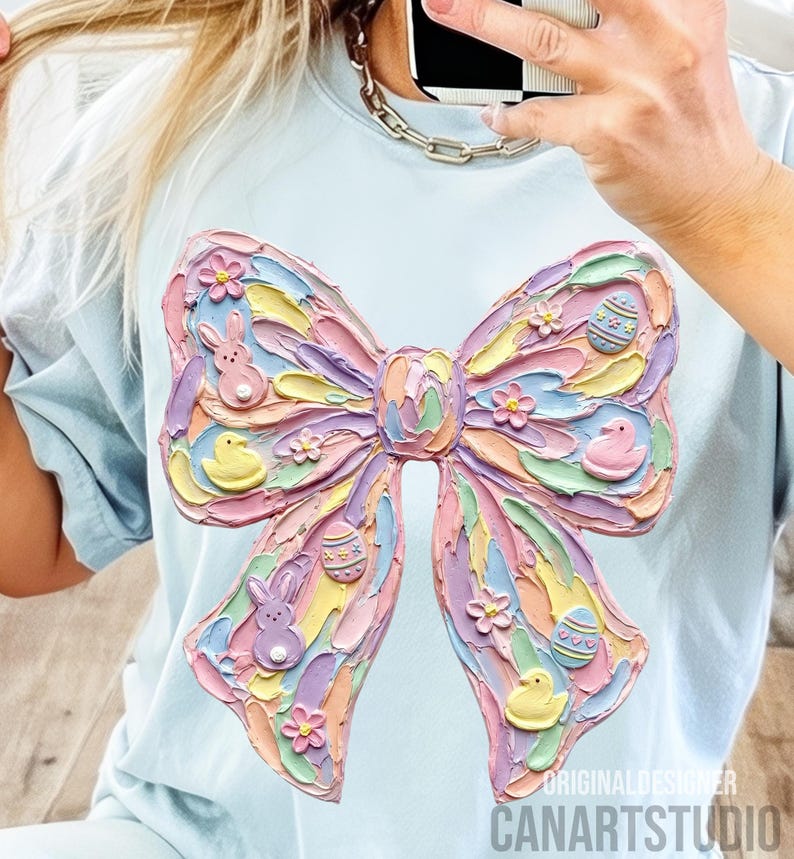 May include: Light blue t-shirt featuring a large, textured bow design in pastel colors. The bow is decorated with Easter-themed elements like bunnies, eggs, and flowers. The text "ORIGINAL DESIGNER CANARTSTUDIO" is visible at the bottom.