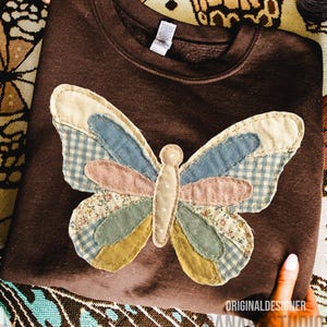 May include: A brown sweatshirt with a large, appliqu&eacute;d butterfly design. The butterfly features patchwork wings in various colors and patterns, including blue gingham, floral prints, and solid pastels. The sweatshirt has a crew neck and long sleeves.