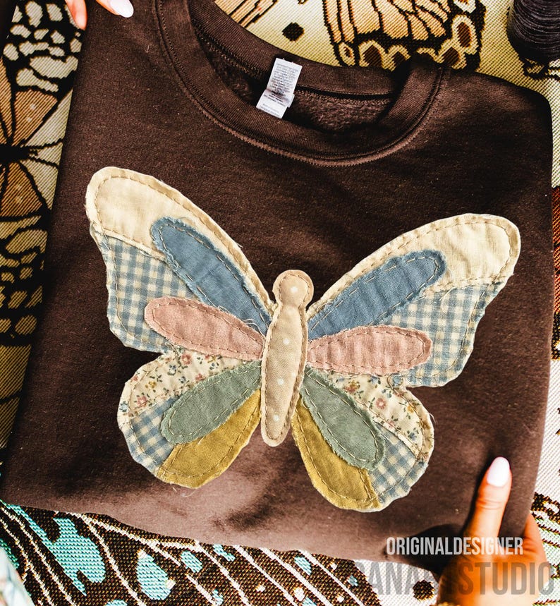 May include: A brown sweatshirt with a large, appliqu&eacute;d butterfly design. The butterfly features patchwork wings in various colors and patterns, including blue gingham, floral prints, and solid pastels. The sweatshirt has a crew neck and long sleeves.