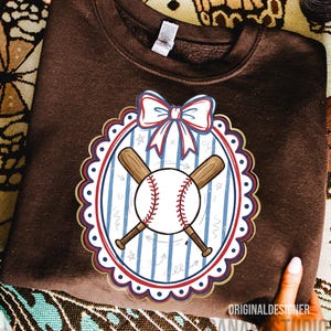 May include: A brown sweatshirt featuring a baseball-themed design. The design includes a baseball, crossed bats, and a bow, all within a decorative frame. The text "ORIGINAL DESIGNER" and "ANDY STUDIO" is visible.