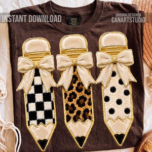 Könnte beinhalten: Ein dunkelbraunes T-Shirt mit drei gestickten Bleistiftmotiven und cremefarbenen Samtschleifen. Die Bleistifte zeigen ein schwarz-weißes Schachbrettmuster, ein braunes Leopardenmuster und schwarze Punkte auf weißem Grund.