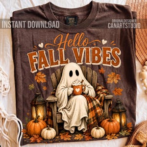 May include: A brown t-shirt featuring a graphic of a ghost sitting in a wooden chair, holding a mug with a heart. The text reads Hello FALL VIBES. The design includes pumpkins, lanterns, and autumn leaves, with a plaid blanket draped over the chair.