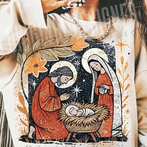 May include: A cream-colored sweatshirt featuring a graphic illustration of the Nativity scene. The design includes Joseph, Mary, and baby Jesus in a manger, with a starry night background and floral accents in orange and gold.