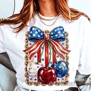 May include: White t-shirt featuring a patriotic design with a bow and cherries. The bow is blue with white stars, and the cherries are red, white, and blue. The design is framed with a leopard print border.