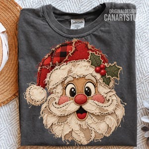 Peut inclure: Un t-shirt gris anthracite orné d'une grande application texturée représentant un visage de Père Noël. Le motif comprend un bonnet à carreaux rouge et noir, une barbe beige effilochée, un nez rouge et du houx vert avec des baies rouges.