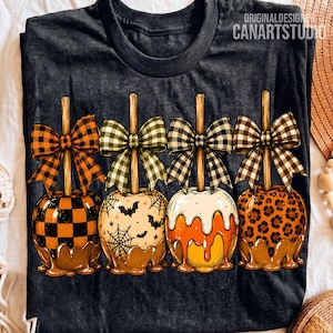 May include: A dark gray t-shirt featuring a graphic of four caramel apples on sticks, each topped with a different patterned bow. The apples display orange and black checkerboard, spiderweb and bat, candy corn, and leopard print designs.
