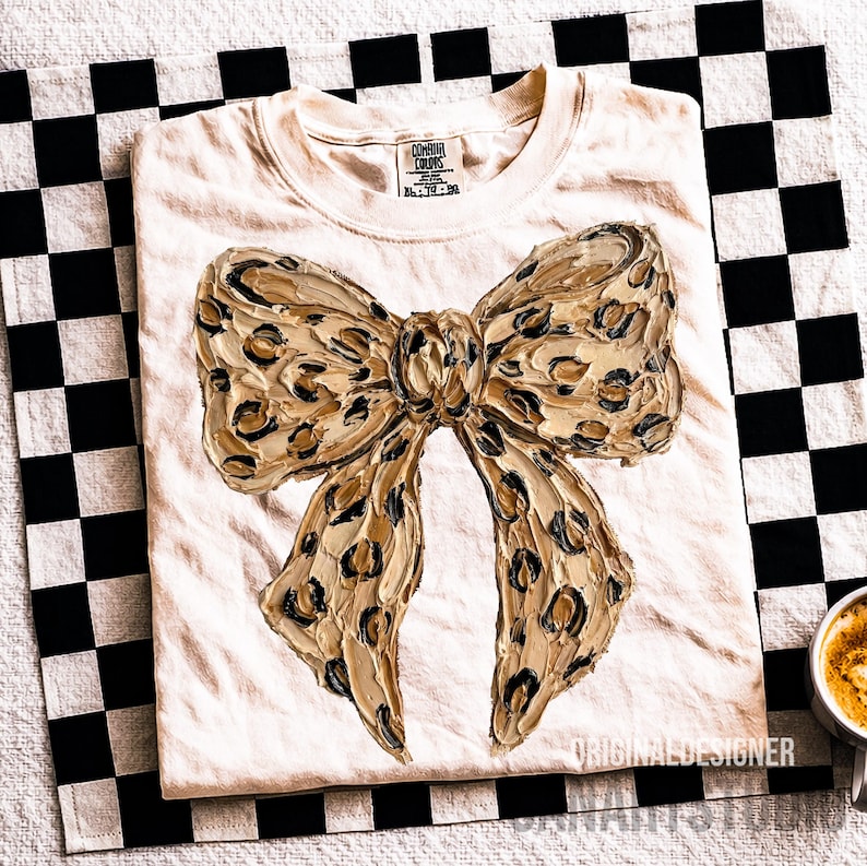 May include: Cream-colored t-shirt featuring a large, textured bow design with a leopard print pattern in shades of tan, brown, and black. The shirt is folded on a black and white checkered surface. The tag reads "COMFORT COLORS".