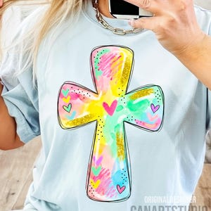 Op de afbeelding: Lichtblauw T-shirt met een kleurrijk kruisontwerp. Het kruis is versierd met aquarel-achtige streken in geel, roze, groen en blauw, geaccentueerd met gouden glitter en kleine hartjes. De tekst "CANARTSTUDIO" is zichtbaar aan de onderkant.