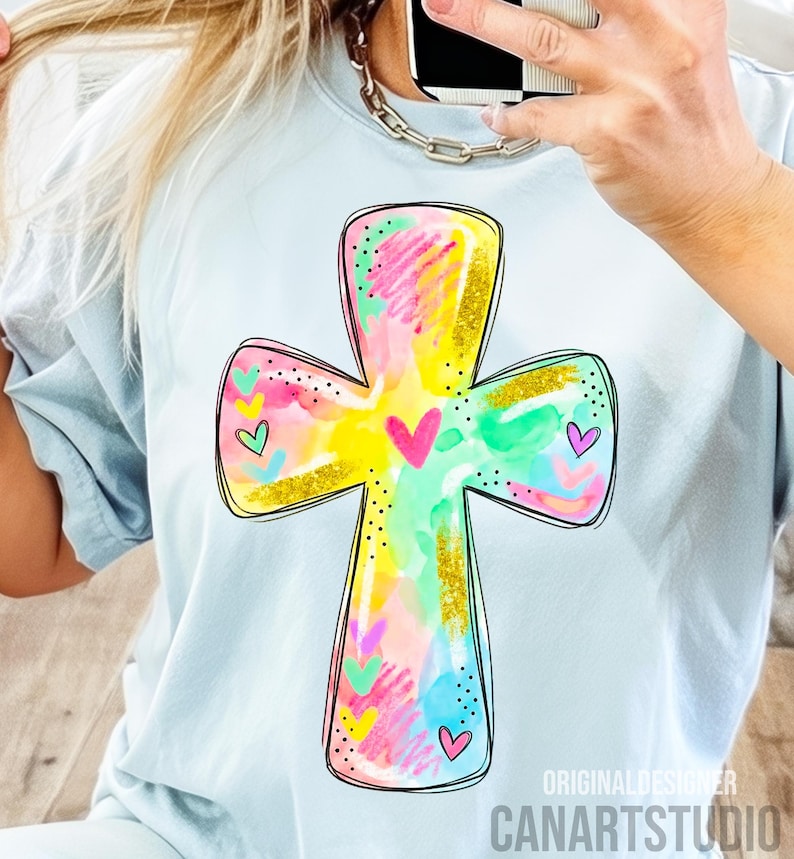 Op de afbeelding: Lichtblauw T-shirt met een kleurrijk kruisontwerp. Het kruis is versierd met aquarel-achtige streken in geel, roze, groen en blauw, geaccentueerd met gouden glitter en kleine hartjes. De tekst "CANARTSTUDIO" is zichtbaar aan de onderkant.