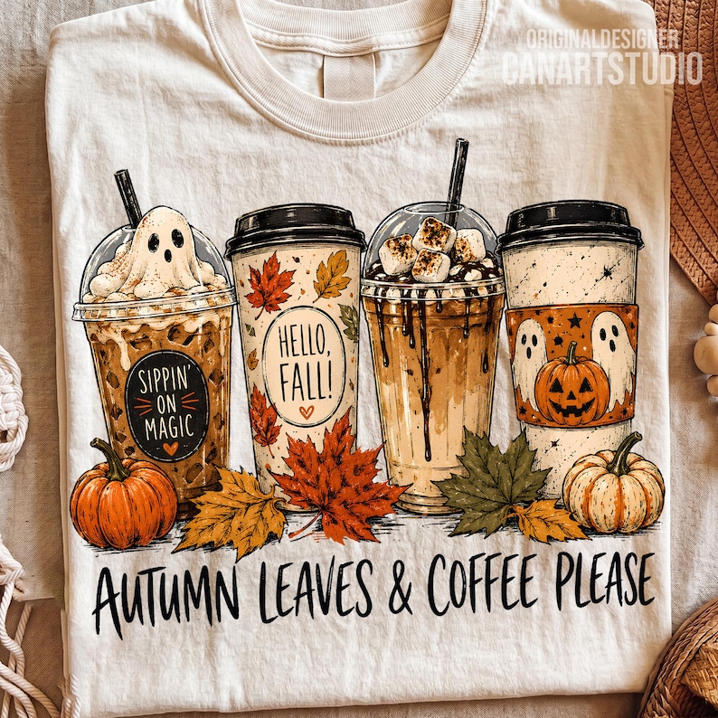 May include: A cream-colored t-shirt featuring a graphic of four coffee cups with autumn themes, including a ghost, pumpkins, and maple leaves. The text reads "Sippin' on Magic," "Hello, Fall!," and "Autumn Leaves & Coffee Please" in black lettering.