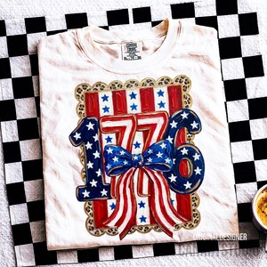 May include: Cream-colored t-shirt with a patriotic design featuring the number "1776" in blue and white stars, a red and white striped bow, and a leopard print border. The shirt is folded on a black and white checkered surface.