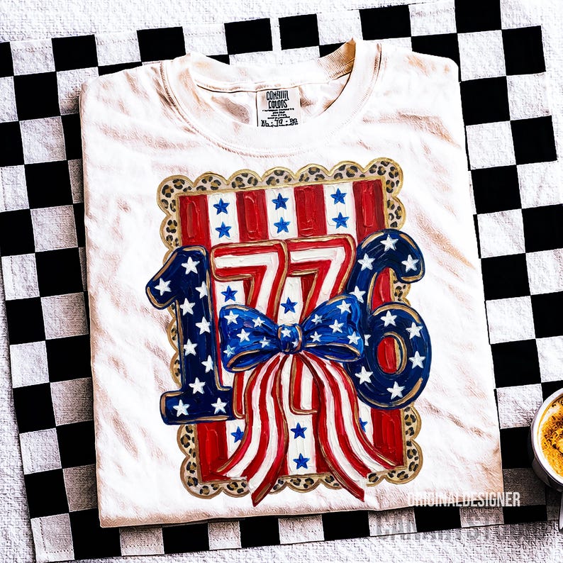 May include: Cream-colored t-shirt with a patriotic design featuring the number "1776" in blue and white stars, a red and white striped bow, and a leopard print border. The shirt is folded on a black and white checkered surface.