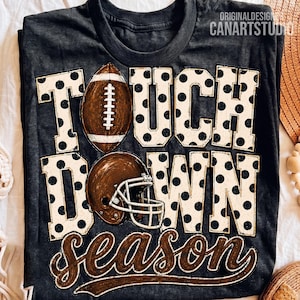 May include: A charcoal grey t-shirt featuring the text TOUCHDOWN SEASON in large block letters with a black polka dot pattern. A brown football replaces the letter O, and a brown football helmet is positioned below it on the shirt.