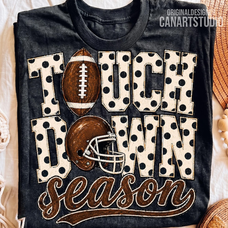 May include: A charcoal grey t-shirt featuring the text TOUCHDOWN SEASON in large block letters with a black polka dot pattern. A brown football replaces the letter O, and a brown football helmet is positioned below it on the shirt.