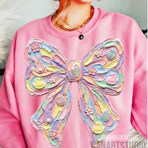 May include: Pink sweatshirt featuring a large, textured bow design with pastel colors. The bow includes Easter-themed elements like bunnies, ducklings, and decorated eggs. The text "ORIGINALDESIGNER SANARTSTUDIO" is visible at the bottom right.