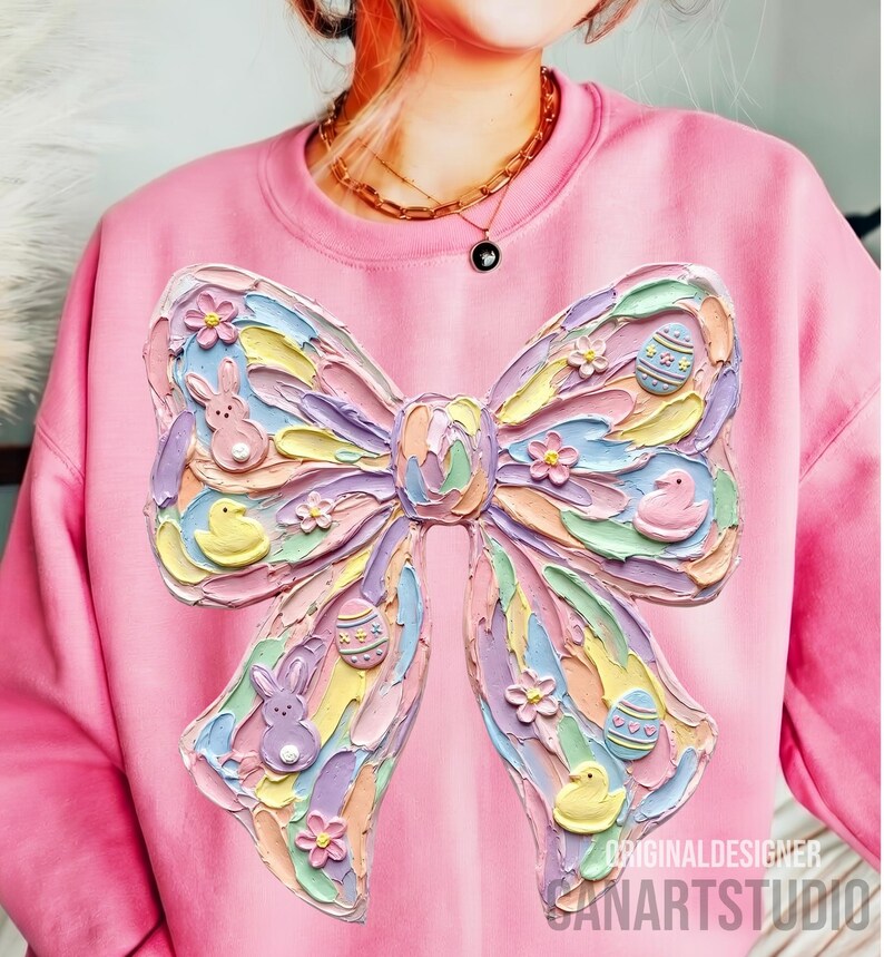 May include: Pink sweatshirt featuring a large, textured bow design with pastel colors. The bow includes Easter-themed elements like bunnies, ducklings, and decorated eggs. The text "ORIGINALDESIGNER SANARTSTUDIO" is visible at the bottom right.