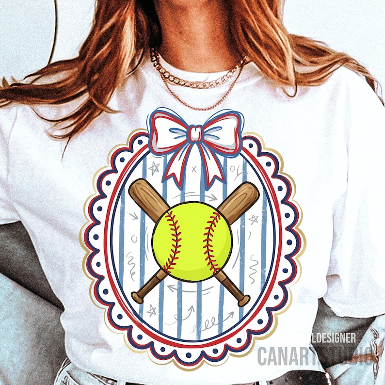 May include: White t-shirt featuring a softball design. The design includes a yellow softball with red stitching, crossed wooden bats, and a decorative oval frame with a red, white, and blue bow. The background has blue stripes and stars.
