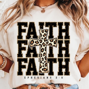 May include: White t-shirt with a black and gold graphic of the word "FAITH" repeated three times with a leopard print cross in the center. The text "EPHESIANS 2:8" is printed below the graphic.
