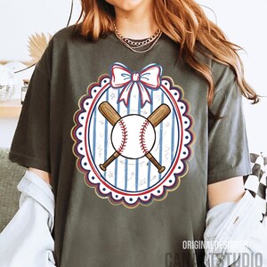 May include: Dark grey t-shirt featuring a baseball design. The design includes a baseball, crossed bats, and a bow in red, white, and blue. The design is inside a scalloped oval with a striped background. Text at the bottom reads "ORIGINAL DESIGNER CANA STUDIO".