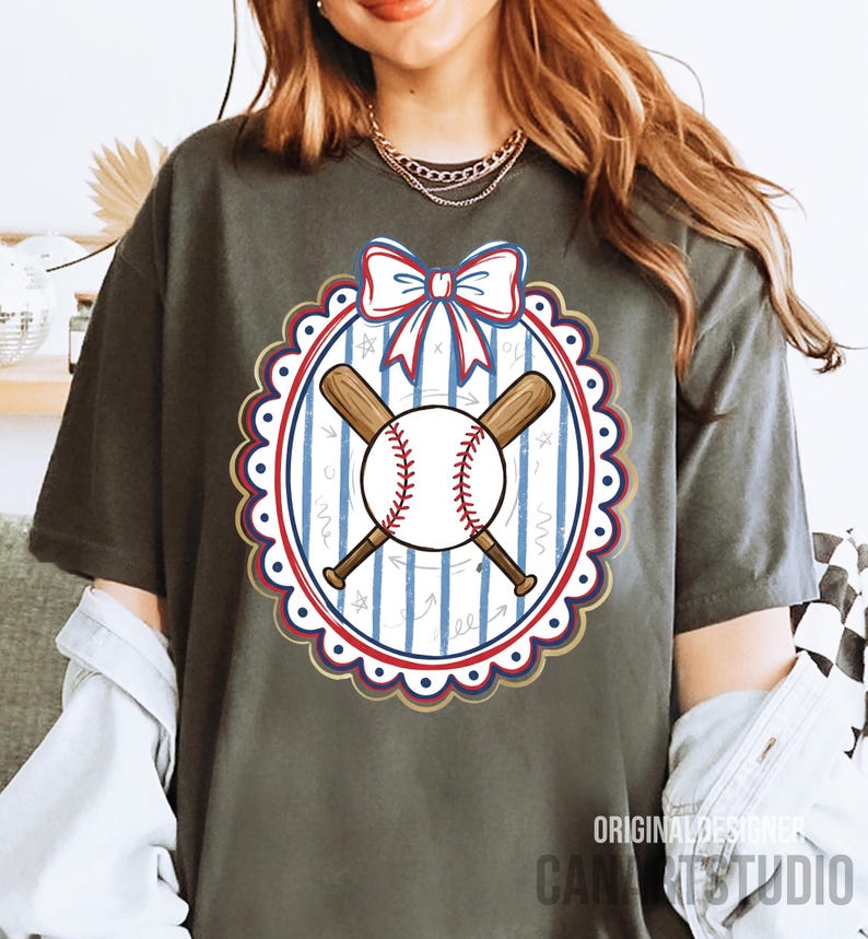 May include: Dark grey t-shirt featuring a baseball design. The design includes a baseball, crossed bats, and a bow in red, white, and blue. The design is inside a scalloped oval with a striped background. Text at the bottom reads "ORIGINAL DESIGNER CANA STUDIO".