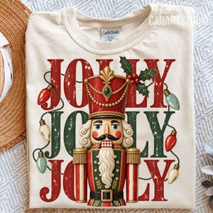 May include: A cream-colored t-shirt featuring a graphic of a nutcracker in a red and green uniform. The text JOLLY is repeated three times in a distressed font. A string of colorful Christmas lights and a sprig of holly decorate the design.