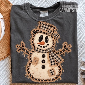 Peut inclure: Un t-shirt gris anthracite avec une application centrale représentant un bonhomme de neige portant un chapeau et une écharpe à carreaux noirs et blancs, un nez en carotte orange et des boutons noirs. Le motif présente des bords en toile de jute effilochés.