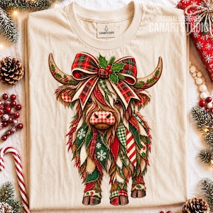 以下が含まれることがあります： 長い角を持つハイランド牛のグラフィックが描かれたクリーム色のTシャツ。ヒイラギの葉が付いた大きな赤と緑のチェック柄のリボンを付けています。牛の鼻は赤と白のチェック柄で、体毛は様々な祝祭的な布のパターンで構成されています。