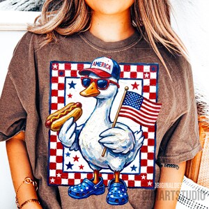 May include: Brown t-shirt featuring a patriotic cartoon duck wearing sunglasses, a baseball cap with "America" on it, and blue star-studded clogs. The duck holds a hot dog and an American flag against a red, white, and blue checkered background.