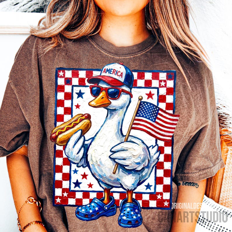 May include: Brown t-shirt featuring a patriotic cartoon duck wearing sunglasses, a baseball cap with "America" on it, and blue star-studded clogs. The duck holds a hot dog and an American flag against a red, white, and blue checkered background.