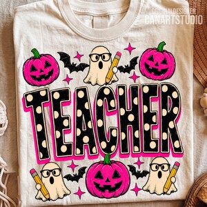 May include: A cream-coloured t-shirt featuring a graphic print with the word TEACHER in large black letters with white polka dots. The design includes three pink jack-o'-lanterns, three ghosts wearing black glasses and holding yellow pencils, and black bats.