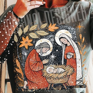 May include: A dark green long-sleeve shirt featuring a religious illustration of the Nativity scene. The design includes Joseph, Mary, and baby Jesus in a manger, with a starry night background and gold accents.