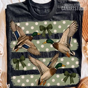 Pu&ograve; includere: Una t-shirt grigio antracite con tre germani reali in volo, fiocchi verdi e uno sfondo a strisce orizzontali verde salvia con pois bianchi. Il testo ORIGINALDESIGN CANARTSTUDIO &egrave; stampato in bianco nell'angolo in alto a destra.