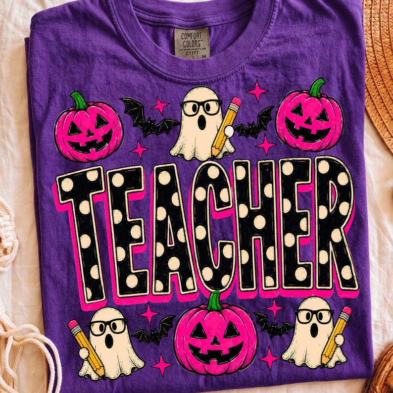 May include: A purple t-shirt featuring a graphic print with the word TEACHER in large black letters with white polka dots. The design includes three pink jack-o'-lanterns, three white ghosts wearing black glasses and holding yellow pencils, and bats.