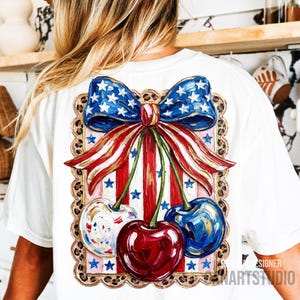 May include: White t-shirt featuring a patriotic design. The design includes a blue bow with white stars, red and white striped ribbons, and three cherries. The cherries are painted in red, blue, and white with gold accents. The design is framed with a leopard print border.